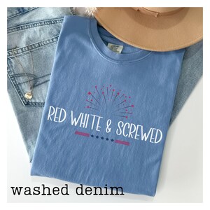 May include: A light blue t-shirt with the text "RED WHITE & SCREWED" in white, featuring a firework design. The shirt is paired with light wash denim jeans and a tan hat. The words "washed denim" are at the bottom.