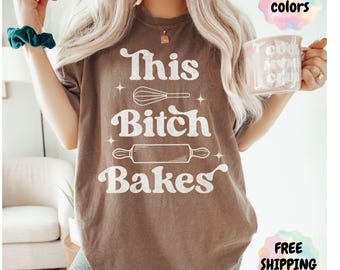 May include: Espresso-colored t-shirt with the text "This Bitch Bakes" in white, featuring a whisk and rolling pin graphic. The shirt is a comfort color and is a casual wear item.