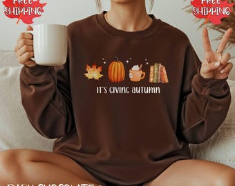 May include: Dark chocolate brown sweatshirt with an autumn-themed graphic. The graphic includes a maple leaf, pumpkin, coffee cup, and books, with the text "It's giving autumn".