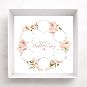 May include: A white box with a floral design for Mother's Day. The design features a wreath of pink peonies and green leaves surrounding six circular spaces. The words "Happy Mother's Day" are written in a script font, with a small heart below.