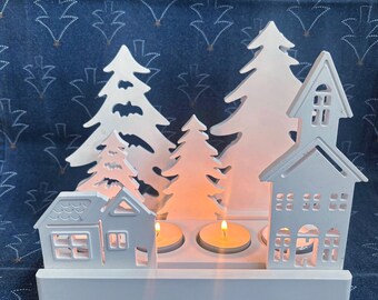 Puede incluir: Portavelas navideño de madera blanca con siluetas recortadas de casas y pinos. El portavelas tiene espacio para tres velas de té, con dos velas encendidas visibles. El fondo es una tela azul con un patrón de árboles repetido.