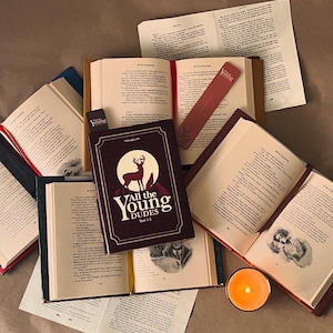 May include: Several open books, including one titled "All the Young Dudes" with a deer illustration. The books have red and blue covers, with bookmarks and a lit candle. The pages are filled with text.
