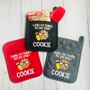 Pode incluir: Três luvas de forno com um design de biscoito e o texto "Obrigado por me tornar inteligente, biscoito". As luvas são vermelhas, pretas e cinzas.