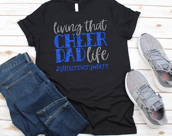 Könnte beinhalten: Schwarzes T-Shirt mit silbernem Glitzertext, der "living that CHEER DAD life #glittereverywhere" lautet. Das Shirt wird mit einer blauen Jeans und grauen Turnschuhen kombiniert.