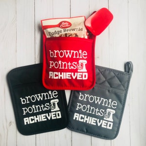 Pode incluir: Três luvas de forno em vermelho, preto e cinza com o texto "brownie points achieved" e um gráfico de uma batedeira.