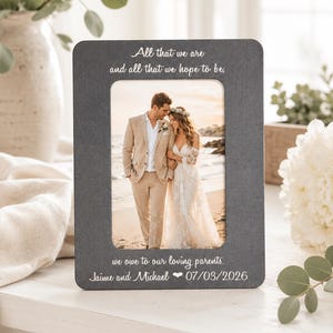 Puede incluir: Un marco de fotos rectangular de color gris oscuro con texto en blanco que dice: All that we are and all that we hope to be, we owe to our loving parents. Jaime and Michael 07/03/2026. El marco contiene una fotografía de una pareja en la playa.