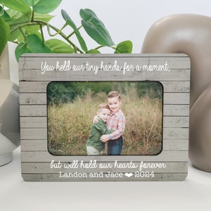 以下が含まれることがあります： 「You held our tiny hands for a moment, but will hold our hearts forever. Landon and Jace 2024」という白い文字が書かれた灰色の木製のフォトフレーム。フレームには、野原で抱き合っている2人の少年の写真が入っています。