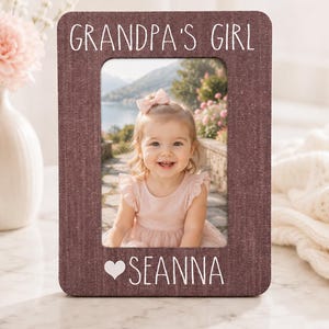May include: A rectangular dark maroon picture frame with a textured finish. The top of the frame features the white text "GRANDPA'S GIRL" and the bottom displays a white heart icon followed by the name "SEANNA" in white capital letters.