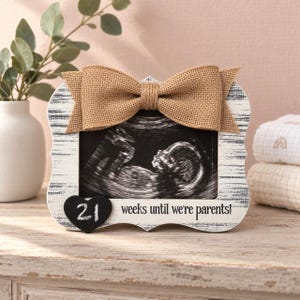 May include: A rectangular picture frame with a distressed white and black wood finish, featuring a tan burlap bow at the top. The frame holds a black and white ultrasound image and includes a black heart-shaped tag with the number 21 and the text 21 weeks until we're parents!
