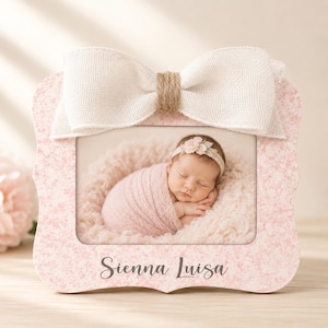 May include: A rectangular photo frame with a light pink floral pattern, featuring a large beige burlap bow at the top. The frame displays a photograph of a sleeping infant wrapped in a pink blanket, and the name Sienna Luisa is printed in dark gray script at the bottom center of the frame.