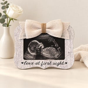 May include: A rectangular picture frame with a white faux-wood grain finish, featuring a beige burlap bow at the top and the black text "love at first sight" with small heart icons at the bottom, displaying a black and white ultrasound image.