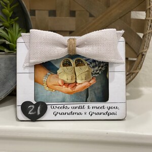 May include: White wooden picture frame with a burlap bow on top. The frame has a photo of a pair of baby shoes in the hands of a couple. The frame also has a black heart with the number 21 written on it and the text "Weeks until I meet you, Grandma & Grandpa!"