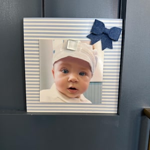 May include: A framed photograph of a baby with blue eyes wearing a white hat. The frame has a blue and white striped pattern and a dark blue bow. The photo is displayed on a dark blue surface.