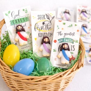 May include: A wicker basket containing green Easter grass, three coloured eggs, and several small Jesus figurines in clear packaging. The packaging has text that reads "God Cares", "Seek First the Kingdom", and "The greatest of these is".