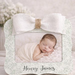 May include: A baby photo frame with a white bow and the name "Henry James" printed on the frame. The frame has a floral pattern and holds a photo of a sleeping infant wrapped in a blanket.