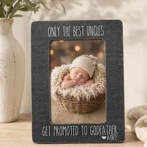 Puede incluir: Un marco de fotos rectangular gris oscuro con el texto "ONLY THE BEST UNCLES GET PROMOTED TO GODFATHER." El marco contiene una foto de un bebé durmiendo en una cesta de mimbre. El marco está sobre una superficie de madera.