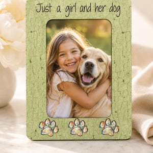 May include: A rectangular picture frame with a light green crackle finish. The top features the black text Just a girl and her dog. The bottom displays three colorful paw print illustrations. A photo of a child hugging a dog is inside.