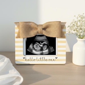 May include: A framed sonogram image with a burlap bow on top. The frame is white with yellow stripes and the text "hello little one". A small white vase with white flowers is to the right.