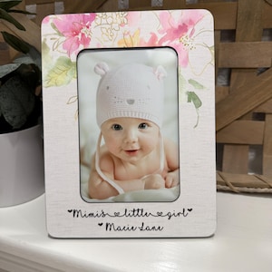 May include: A white wooden picture frame with a watercolor floral design. The frame has a photo of a baby wearing a white hat. The text "Mimi's little girl" and "Marie Lane" is engraved on the frame.