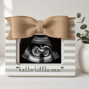 May include: A rectangular picture frame with horizontal sage green and white stripes, featuring a large burlap bow at the top. The frame displays a black and white ultrasound image and the text hello little one in black script with small hearts.