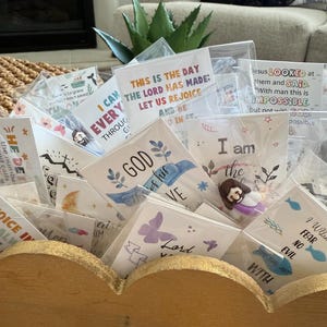 May include: A wooden display box with a gold-painted scalloped edge, filled with small, clear-wrapped cards. The cards feature inspirational quotes and religious imagery, including butterflies, fish, and floral designs. Some cards have text that reads "This is the day the Lord has made."