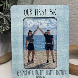 May include: A light blue photo frame with the text "OUR FIRST 5K" and "THE START OF A HEALTHY LIFESTYLE TOGETHER 09.06.25". The frame holds a photo of two people high-fiving on a beach.
