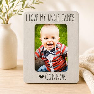 Puede incluir: Un marco de fotos rectangular gris claro con el texto "I LOVE MY UNCLE JAMES" en la parte superior y "CONNOR" en la inferior. El marco contiene una foto de un bebé sonriente con pajarita.