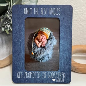 May include: A blue photo frame with the text "ONLY THE BEST UNCLES GET PROMOTED TO GODFATHER" in white. The frame holds a photo of a newborn baby wearing a striped hat. The baby is nestled in a fluffy blue blanket.