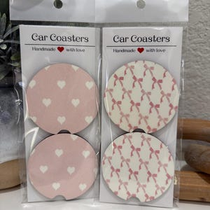 May include: Two sets of car coasters in clear plastic packaging. One set features pink coasters with white hearts, the other with pink bows on a white background. The text "Car Coasters" and "Handmade with love" are printed on the packaging.