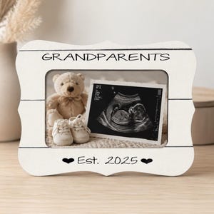 May include: A white picture frame with the word "GRANDPARENTS" at the top and "Est. 2025" at the bottom. The frame holds a sonogram image, a teddy bear, and baby shoes.