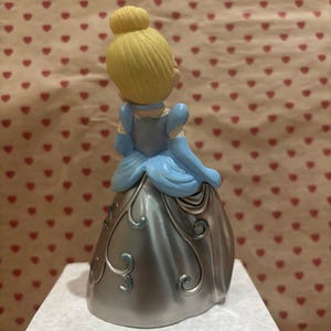 May include: A figurine of a princess, viewed from the back, with blonde hair in a bun and wearing a light blue dress with silver accents. The dress has a metallic sheen and decorative swirls. The background features a pattern of red hearts.