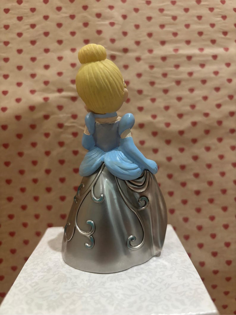 May include: A figurine of a princess, viewed from the back, with blonde hair in a bun and wearing a light blue dress with silver accents. The dress has a metallic sheen and decorative swirls. The background features a pattern of red hearts.