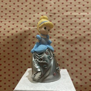 May include: A figurine of Cinderella in a light blue gown with silver accents and a blonde updo. The figurine is standing on a white box with a patterned design. The background is a light brown color with a pattern of red hearts.