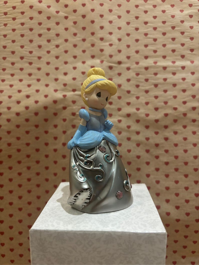 May include: A figurine of Cinderella in a light blue gown with silver accents and a blonde updo. The figurine is standing on a white box with a patterned design. The background is a light brown color with a pattern of red hearts.