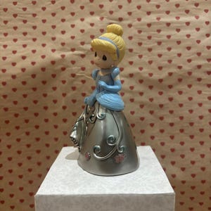 May include: A figurine of a princess in a blue dress with a silver skirt, blonde hair, and a blue headband. The figurine is on a white box with the word "Precious" visible. The background is a pattern of red hearts.