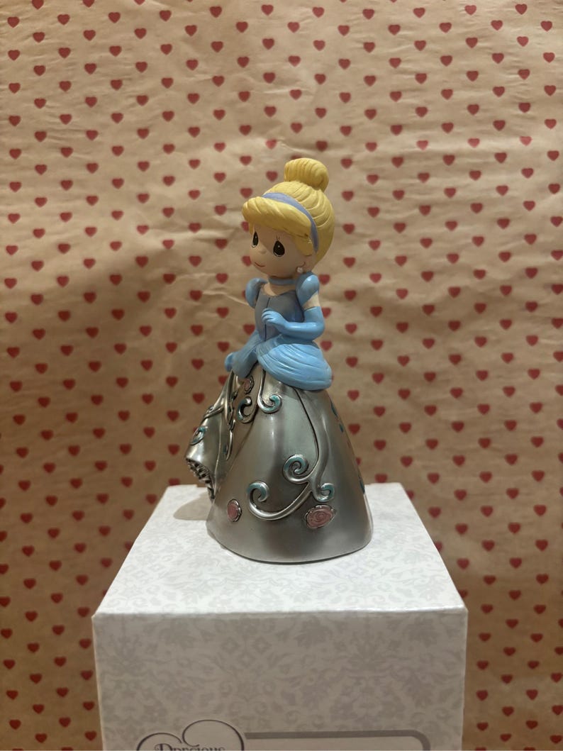 May include: A figurine of a princess in a blue dress with a silver skirt, blonde hair, and a blue headband. The figurine is on a white box with the word "Precious" visible. The background is a pattern of red hearts.