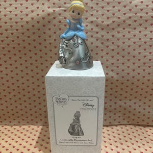 May include: A Precious Moments Cinderella decorative bell figurine. The figurine is hand-painted with a blue dress and blonde hair. The bell is silver with pink floral accents and is displayed on a white box.