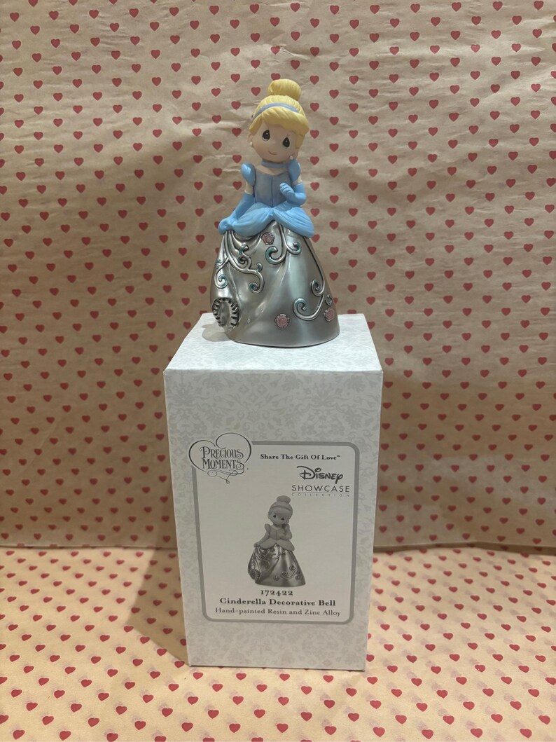 May include: A Precious Moments Cinderella decorative bell figurine. The figurine is hand-painted with a blue dress and blonde hair. The bell is silver with pink floral accents and is displayed on a white box.