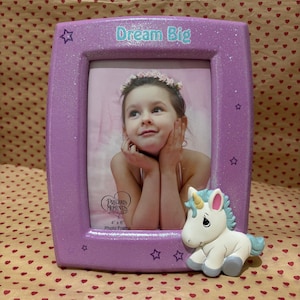 May include: A lavender glitter photo frame with the words "Dream Big" at the top. The frame has a picture of a young girl and a small unicorn figurine on the bottom right. The frame is designed to hold a 4" x 6" photo.
