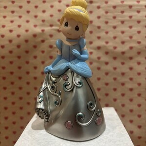 May include: A figurine of Cinderella in a blue dress with a silver skirt, adorned with pink and teal accents. Her blonde hair is styled in a bun. The figurine is on a white box with a patterned design.
