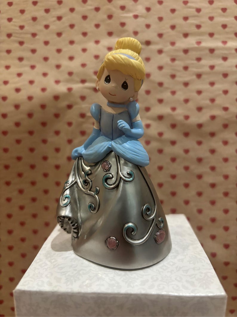 May include: A figurine of Cinderella in a blue dress with a silver skirt, adorned with pink and teal accents. Her blonde hair is styled in a bun. The figurine is on a white box with a patterned design.