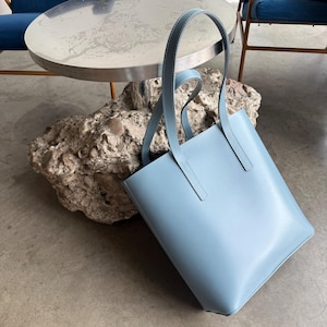 May include: A light blue tote bag with a smooth, matte finish and two long, matching shoulder straps. The bag features clean, vertical lines and visible white stitching along the edges of the straps, creating a structured and simple rectangular shape.