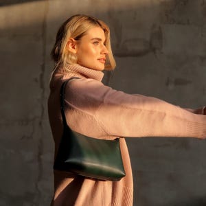 Puede incluir: Un bolso de hombro de cuero verde oscuro con una correa larga. El bolso se sostiene contra una persona que lleva un jersey de cuello alto rosa claro. El fondo es una pared gris neutra.