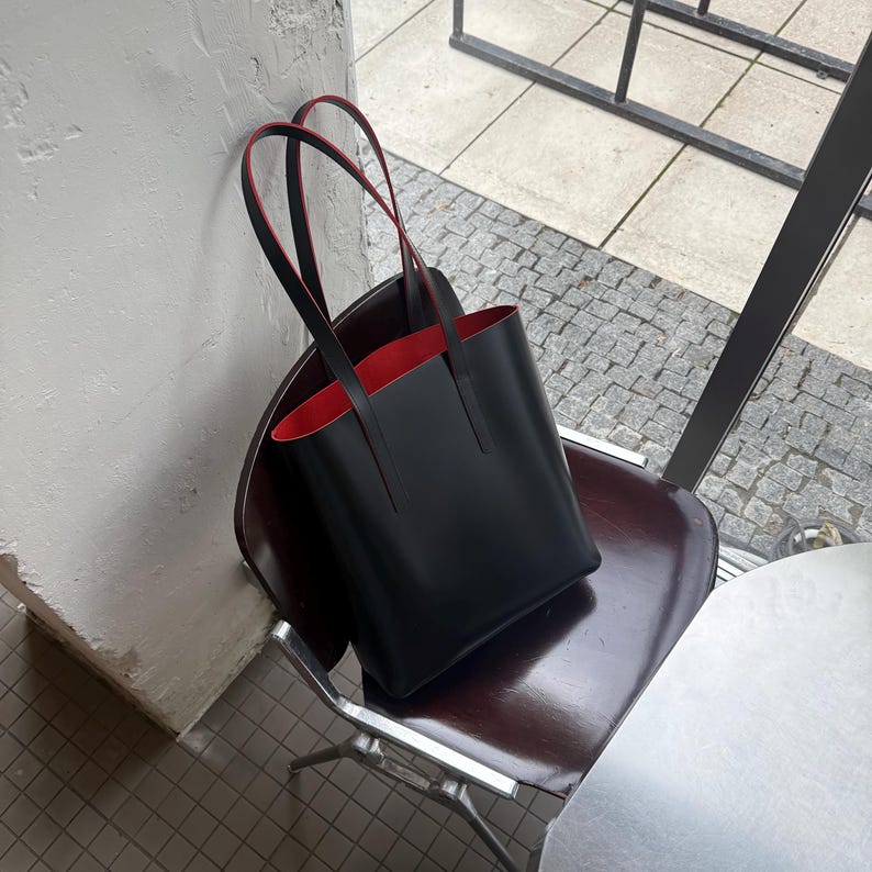 May include: A structured black tote bag with a smooth finish and two long shoulder straps. The interior of the bag is lined with a vibrant red material, which is also visible along the edges of the straps and the top opening of the bag.
