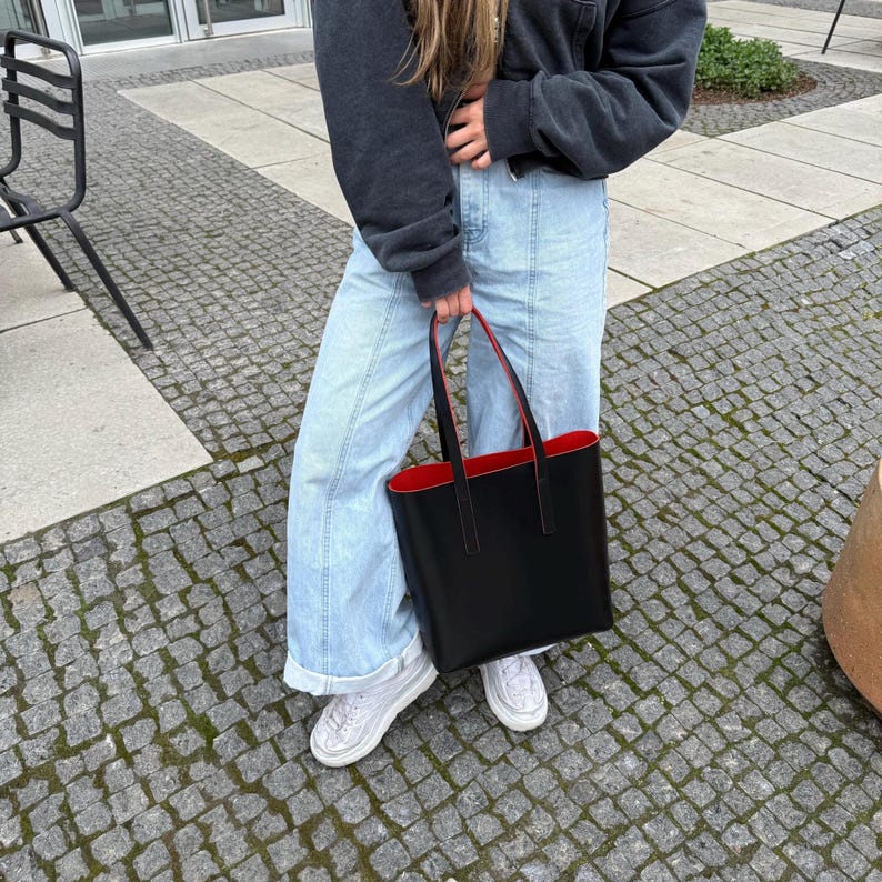 May include: A black tote bag with a vibrant red interior lining and two thin black shoulder straps. The bag features a structured rectangular shape with clean lines and is held by a person wearing a dark gray sweatshirt, light blue jeans, and white sneakers.