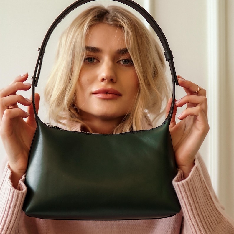 Puede incluir: Un bolso de hombro de cuero verde oscuro con forma curva y una correa larga y fina. El bolso tiene cierre de cremallera y un dise&ntilde;o elegante y minimalista. La correa es de color marr&oacute;n oscuro. El bolso es sostenido por una persona.