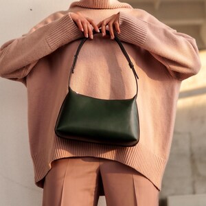 Puede incluir: Un bolso de hombro de cuero verde oscuro con forma curva y correa fina. El bolso se muestra frente a una persona que lleva un su&eacute;ter y pantalones de color rosa claro. El dise&ntilde;o del bolso es simple y elegante.