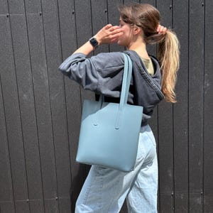 May include: A solid light blue tote bag with a rectangular shape and two vertical shoulder straps. The bag features a smooth, matte finish and a clean, structured design, providing a simple and functional accessory for carrying daily essentials.