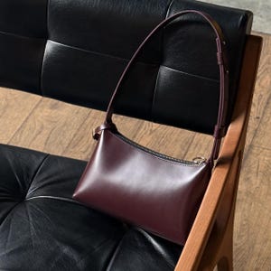 Op de afbeelding: Een bordeauxrode leren schoudertas met een gebogen vorm en een ritssluiting. De tas heeft een dunne riem en een goudkleurige gesp. De tas ligt op een zwarte leren stoel met een houten frame.