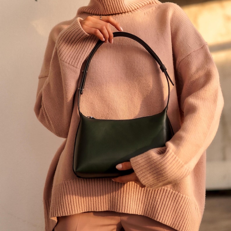 Puede incluir: Un bolso de hombro de cuero verde oscuro con una correa negra. El bolso tiene forma rectangular y cierre de cremallera. La persona lleva un jersey de cuello alto rosa claro.
